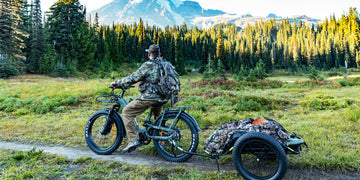 A hunter riding the Himiway D5 2.0 camo fat tire electric bike with a loaded hunting trailer through a mountain meadow.