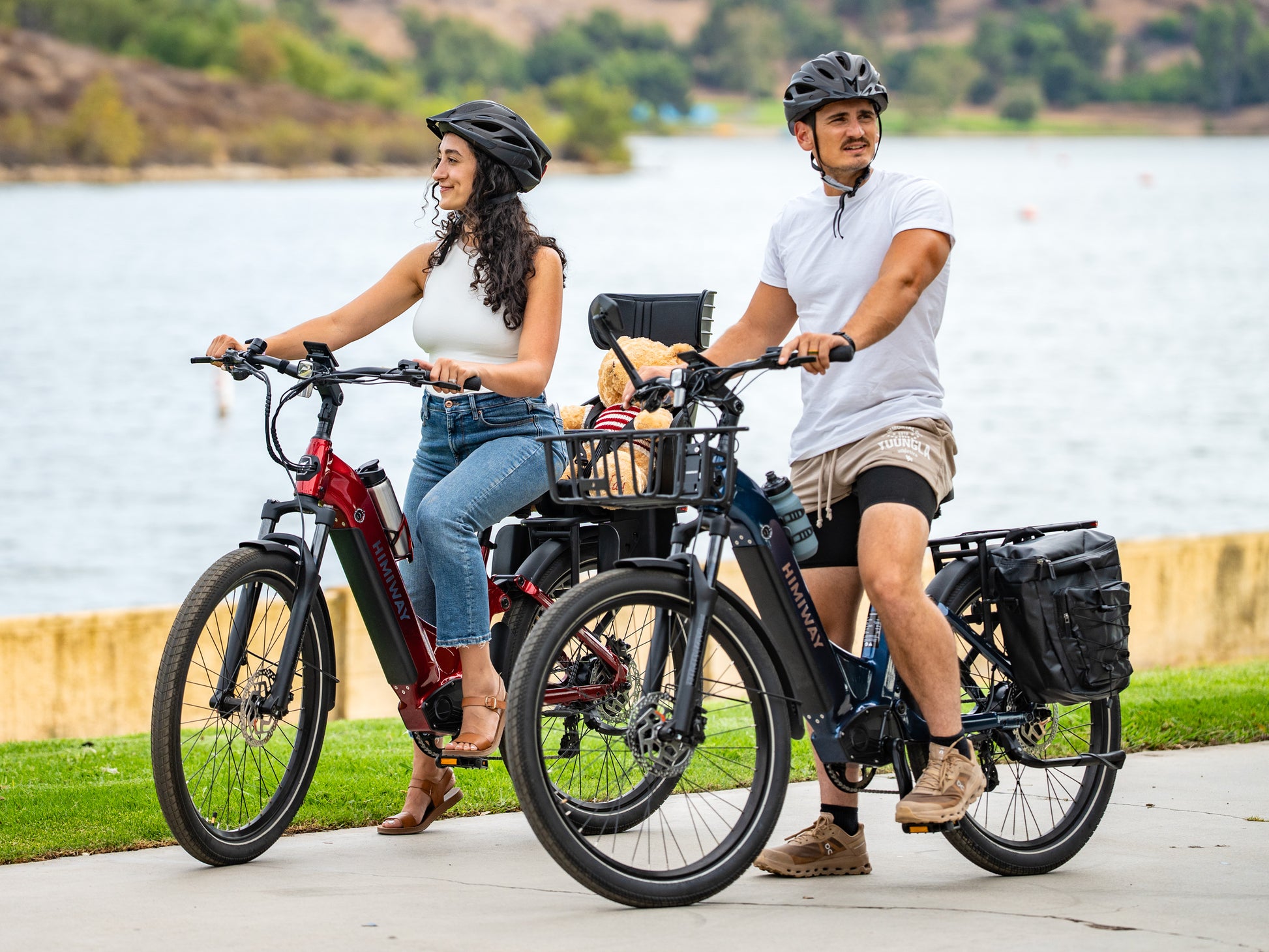 A couple rides red and Midnight Blue Himiway A7 Commuter e-bikes along a lakeside path, the blue bike carrying a teddy bear in its front basket.