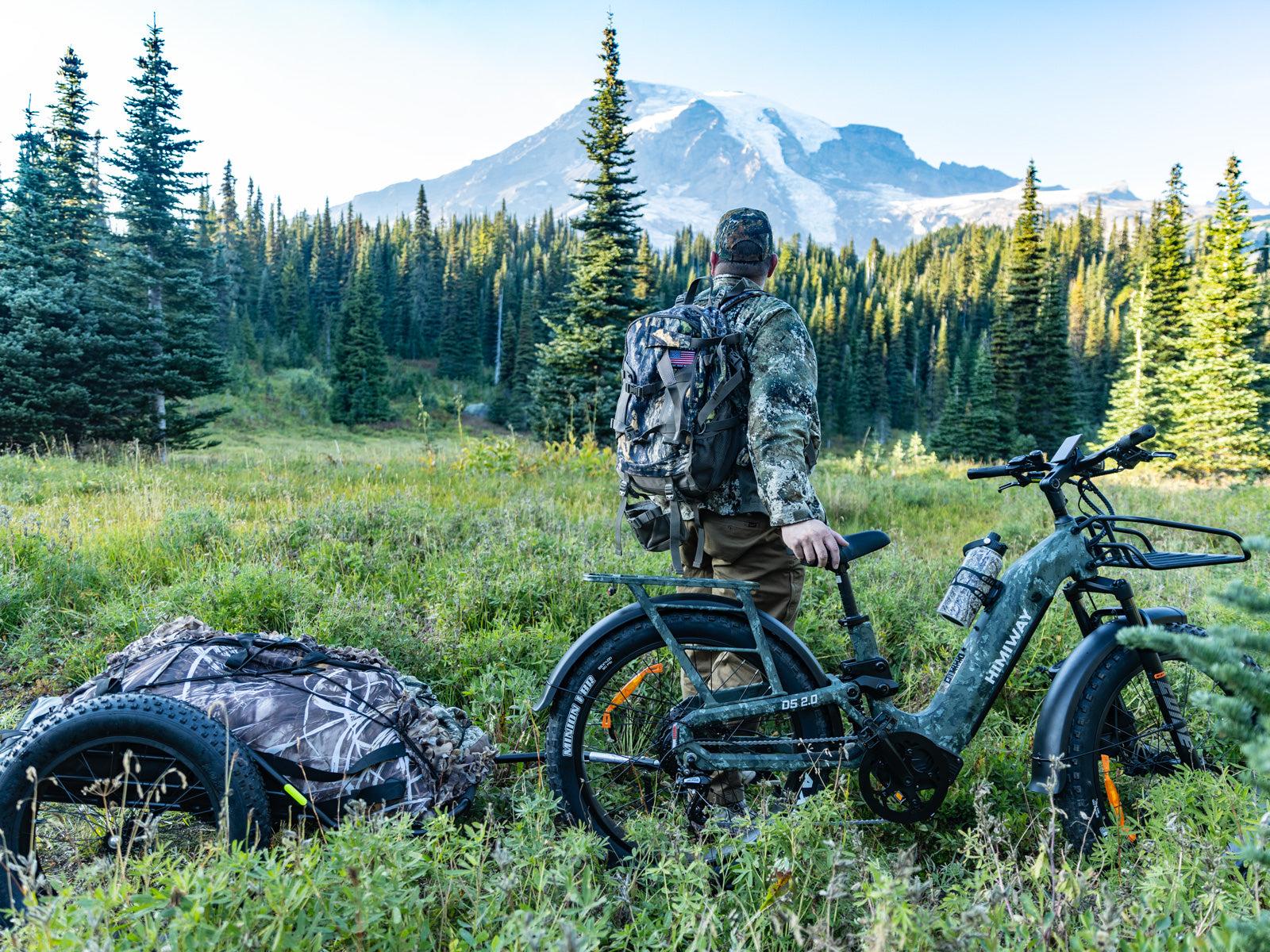 A hunter standing in a mountain meadow next to his Himiway D5 2.0 camo full suspension fat tire electric bike pulling a loaded hunting trailer.