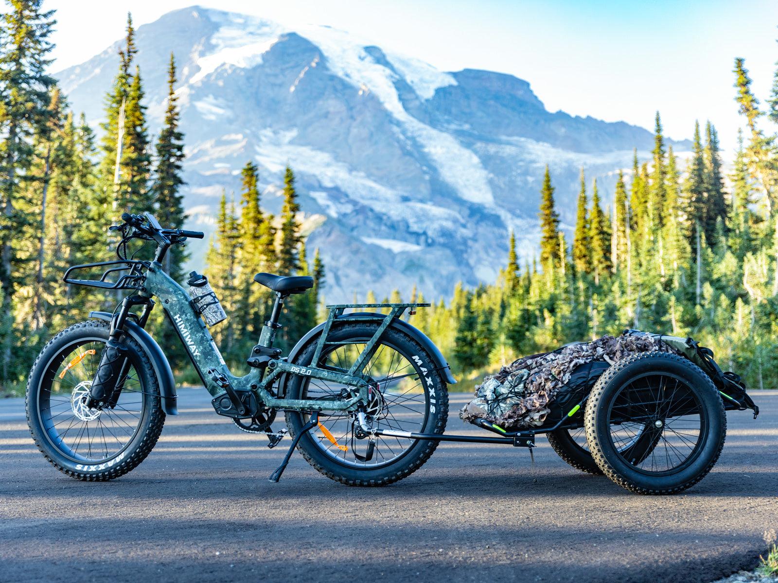 The Himiway D5 2.0 camo fat tire electric bike pulling a loaded hunting trailer with a stunning snow-capped mountain background.