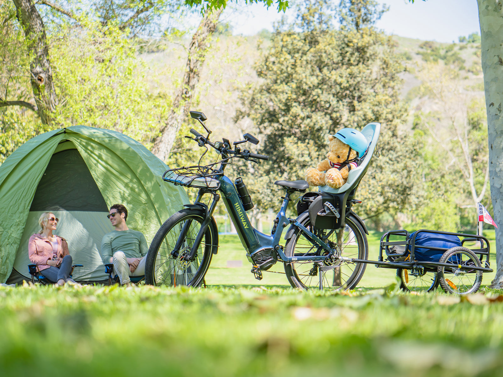 At a sunny campsite, a Himiway A7 Commuter e-bike holds a helmeted teddy in a child seat and tows a loaded trailer while a couple relaxes by their tent.