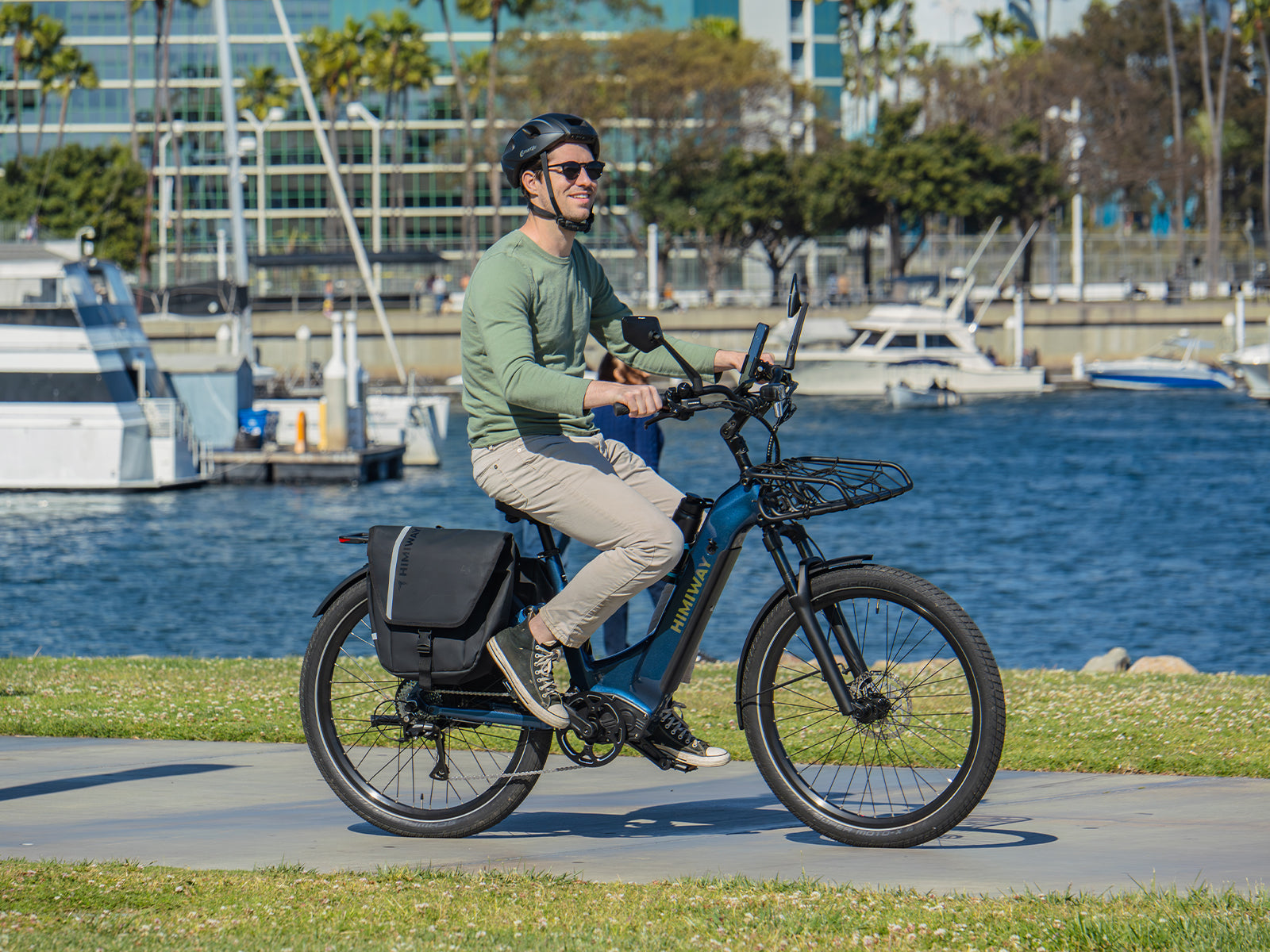 A smiling rider in a helmet cruises a sunny marina path on a Midnight Blue Himiway A7 Commuter e-bike with a front rack and rear pannier