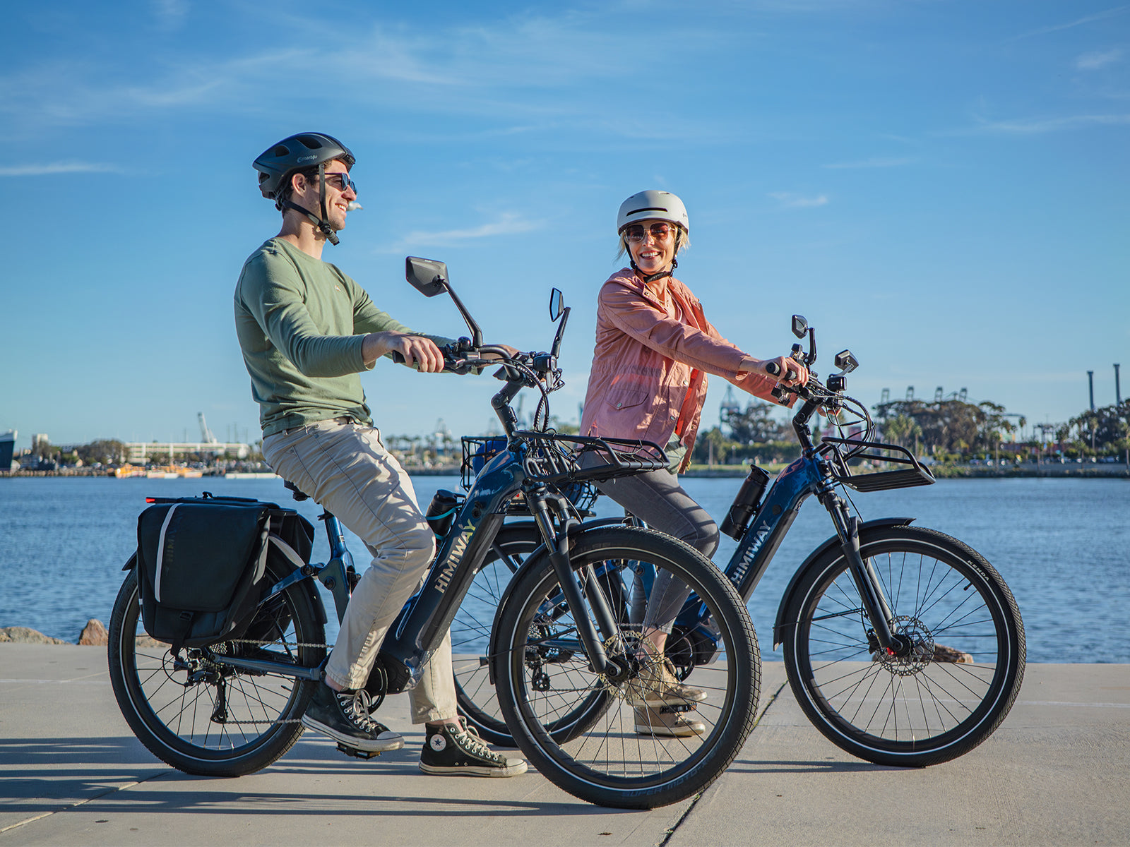 A smiling couple in helmets ride Himiway A7 Commuter e-bikes side by side along a sunny waterfront promenade, one bike loaded with a rear pannier.
