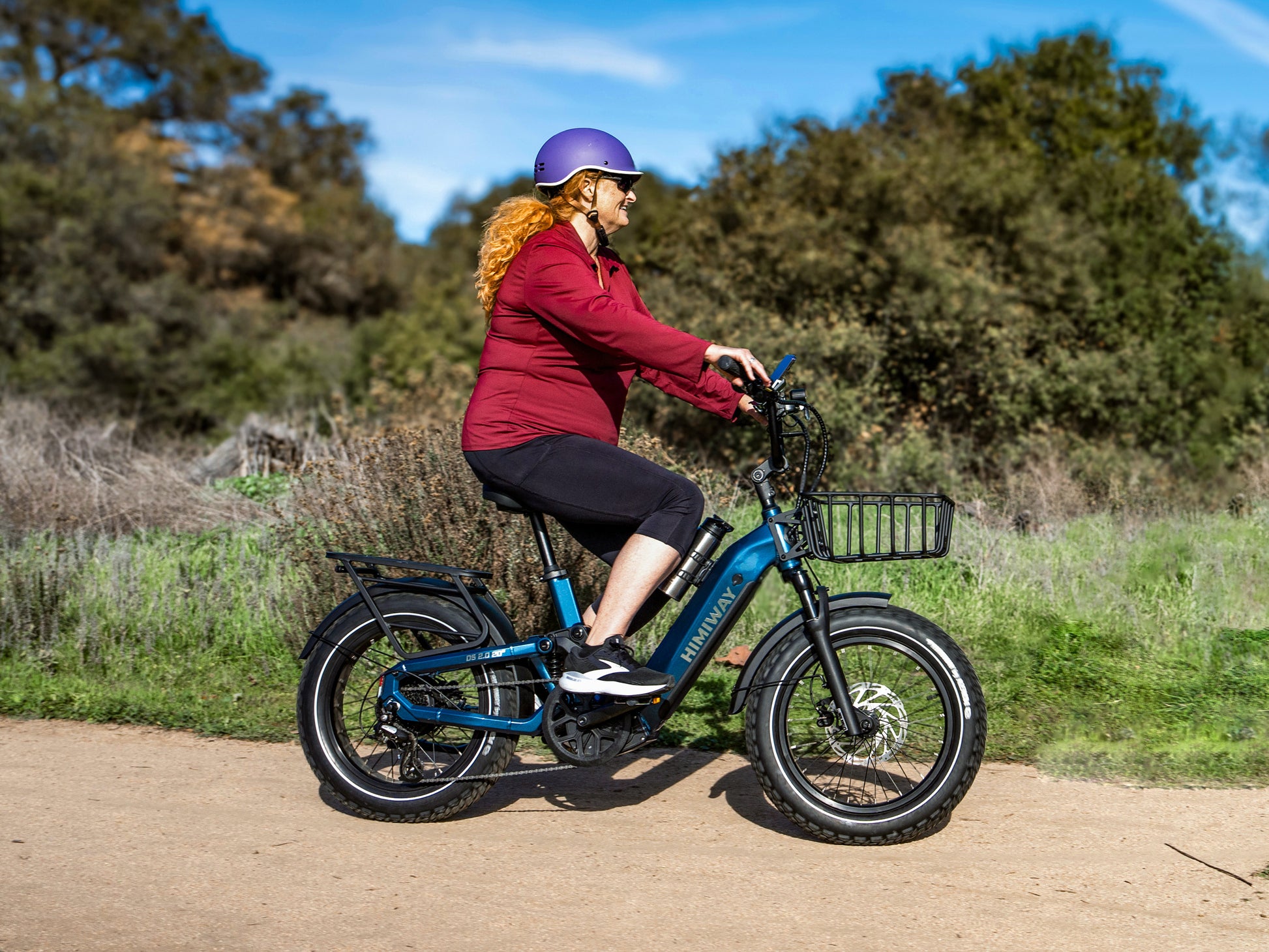 A senior woman riding the blue Himiway D5 2.0 20" step-thru fat tire electric bike on a dirt trail.