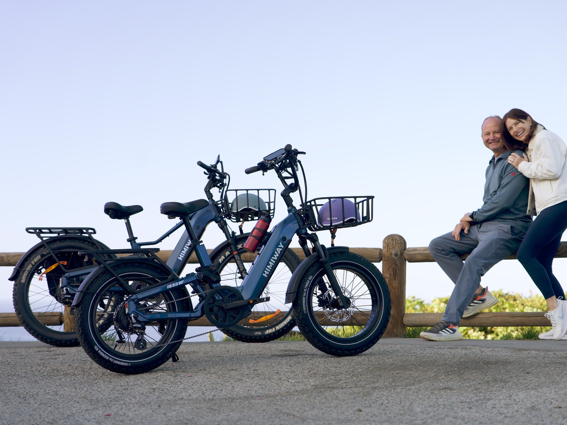 A smiling senior couple resting by a wooden fence with their blue Himiway D5 2.0 20" step-thru fat tire electric bikes.