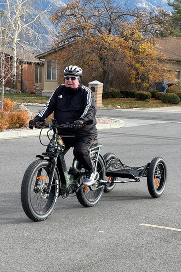 A senior man riding a Himiway D5 2.0 Camo fat tire e-bike towing a cargo trailer on a paved road in a residential neighborhood.