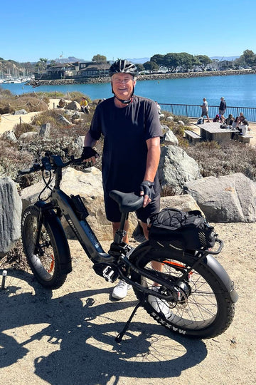 Himiway D5 2.0 Step-Through electric bike with a mature male rider smiling at the camera on a road near blue water and rocks, featuring a rear bag and water bottle holder
