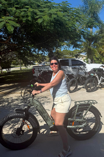 Woman holding a himiway D5 2.0 Camo fat tire full suspension ebike in an outdoor setting with trees and vehicles in the background